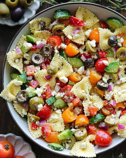 Bowl of bowtie pasta salad with tomatoes, olives, avocado, feta cheese, and vegetables on a wooden table