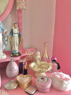 Pink vanity table with perfume bottles, Virgin Mary figurine, gold compact, and feminine accessories