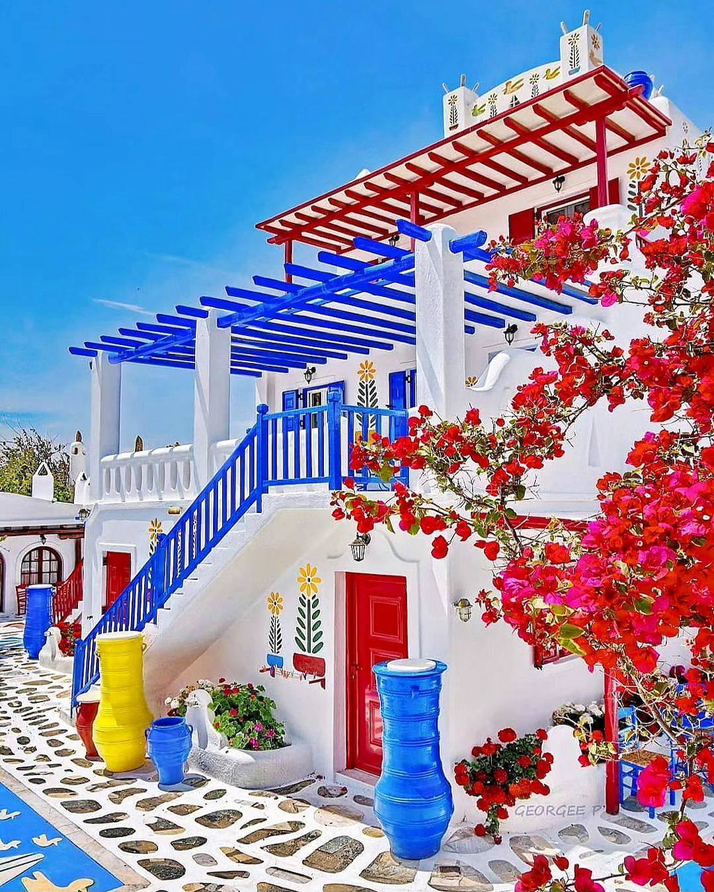 White Greek building with blue pergola, red door, and pink bougainvillea flowers