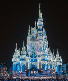 Cinderella Castle lit up at night with blue lights and fireworks, crowds visible at base