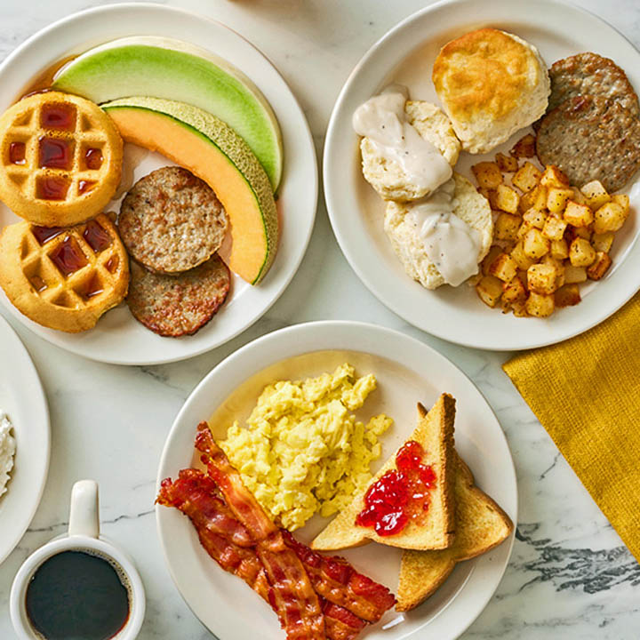 Three white plates with waffles, fruit, scrambled eggs, bacon, sausage, biscuits, and a cup of coffee on marble surface