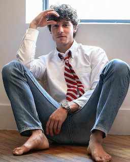 Young man with curly hair in white shirt and red striped tie sitting barefoot on floor