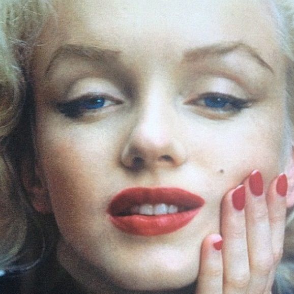 Close-up of a blonde woman with red lipstick and red nails touching her cheek