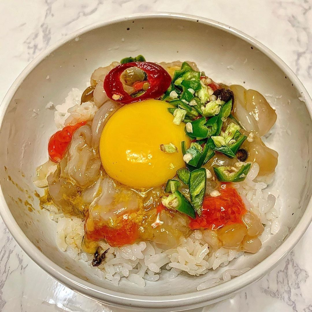 White bowl with rice topped with marinated crab, egg yolk, red pepper, and green onions on marble surface