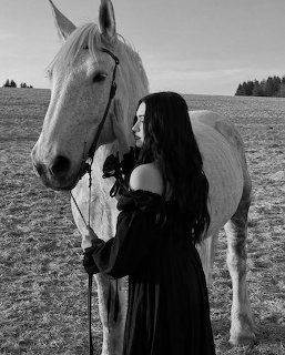 Woman in black dress standing next to white horse in field, black and white photo