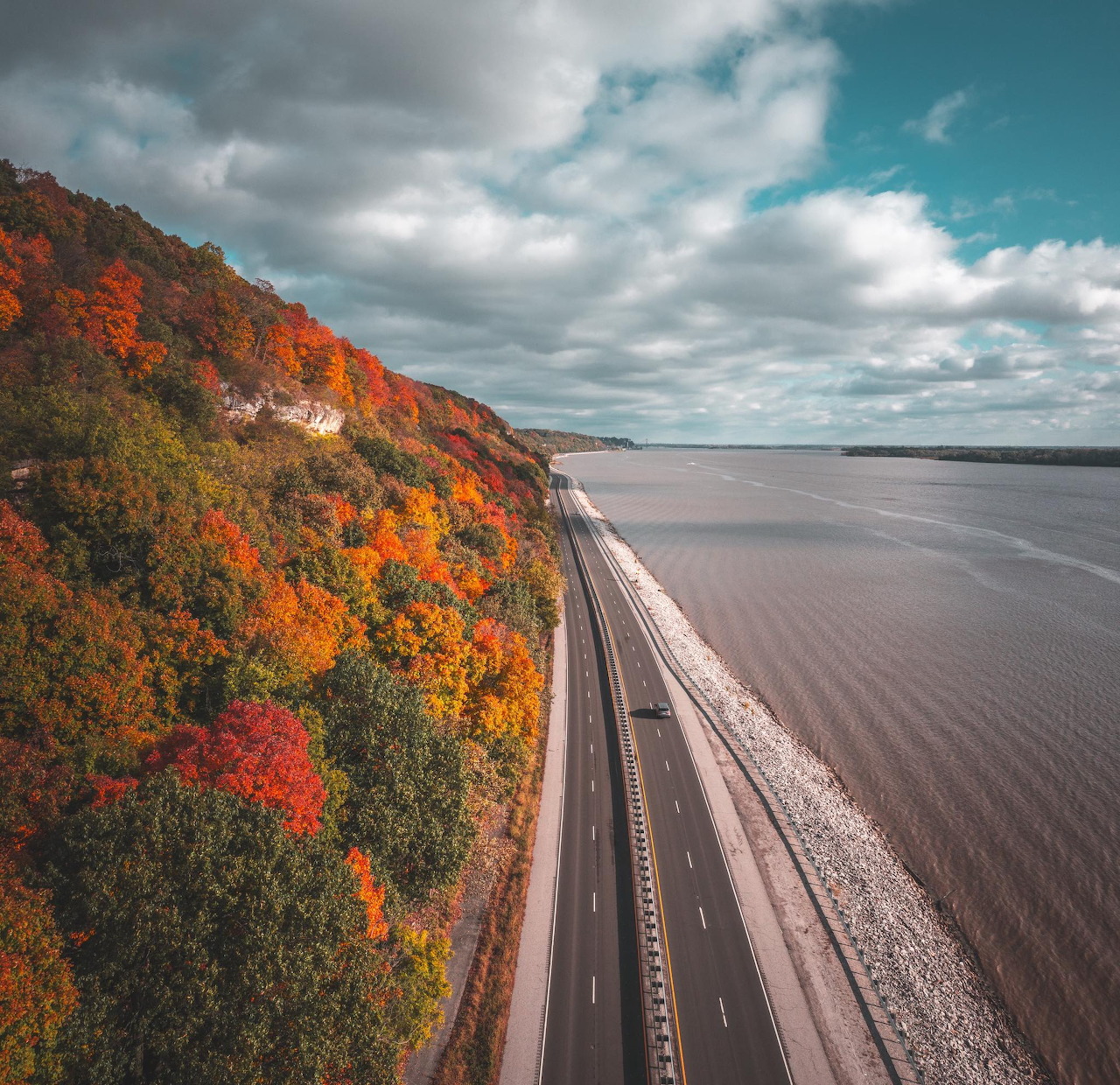 Aerial view of a road along a river beside a hillside covered in colorful autumn trees