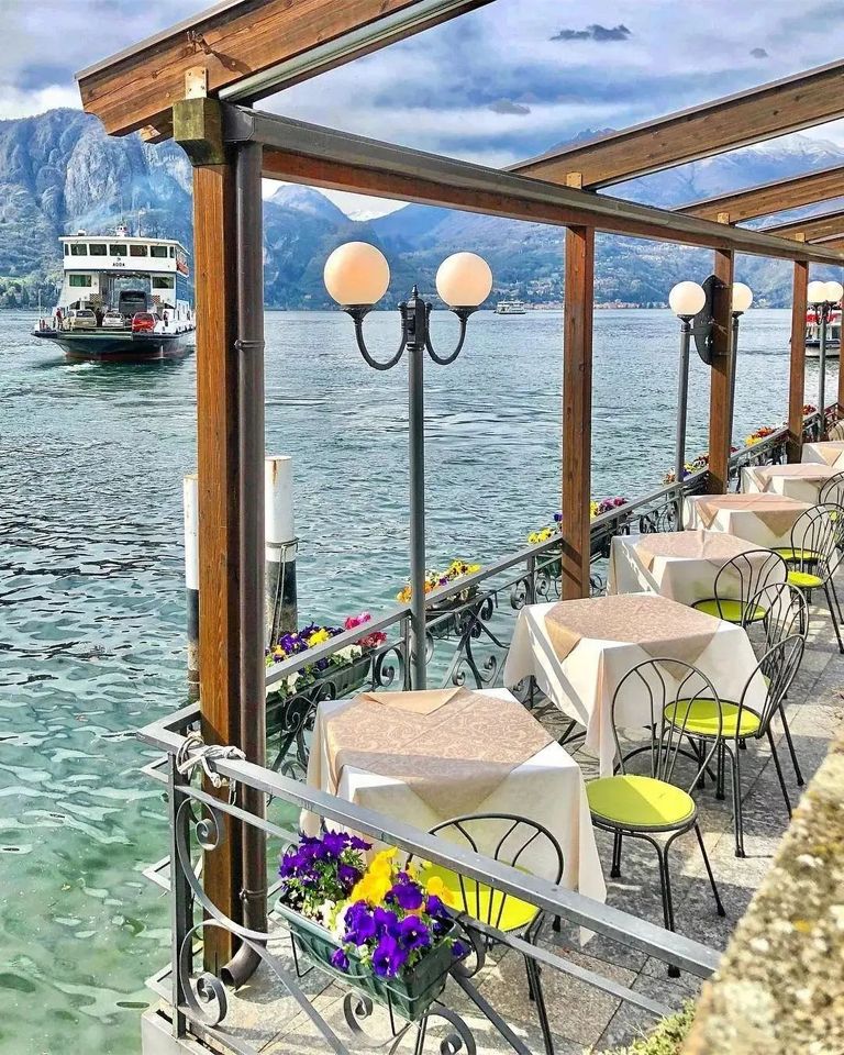Outdoor lakeside restaurant terrace with tables, lamp posts, purple flowers, and a ferry on a blue lake