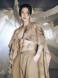 Young Asian man in open beige linen robe and wide trousers posing in a studio with draped fabric backdrop