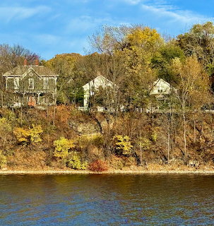 Houses on a tree-covered bluff above a river with autumn foliage and blue water