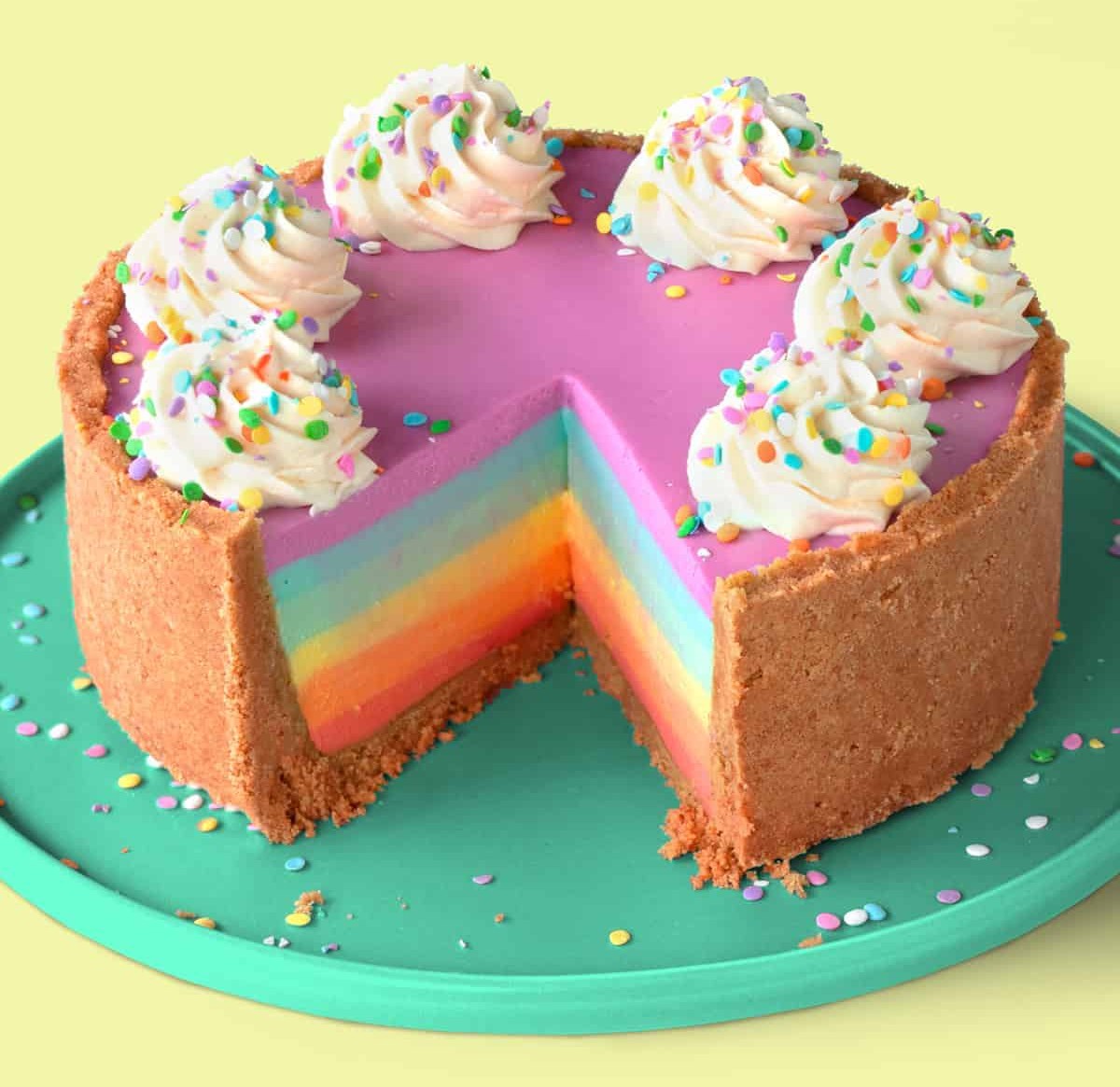 Rainbow-layered cheesecake with pink top, whipped cream swirls, and colorful sprinkles on a teal plate