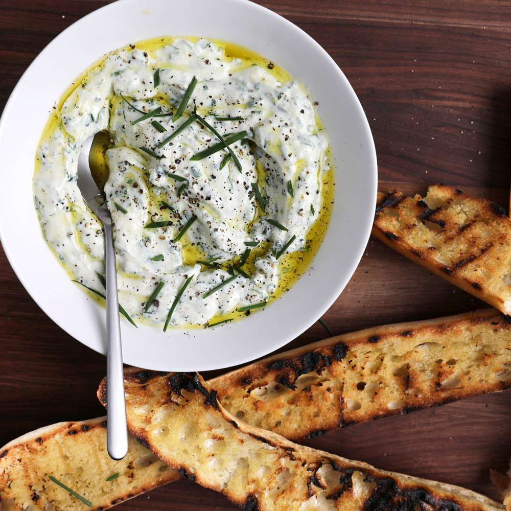 White bowl of labneh dip with herbs and olive oil, accompanied by grilled bread on wooden table