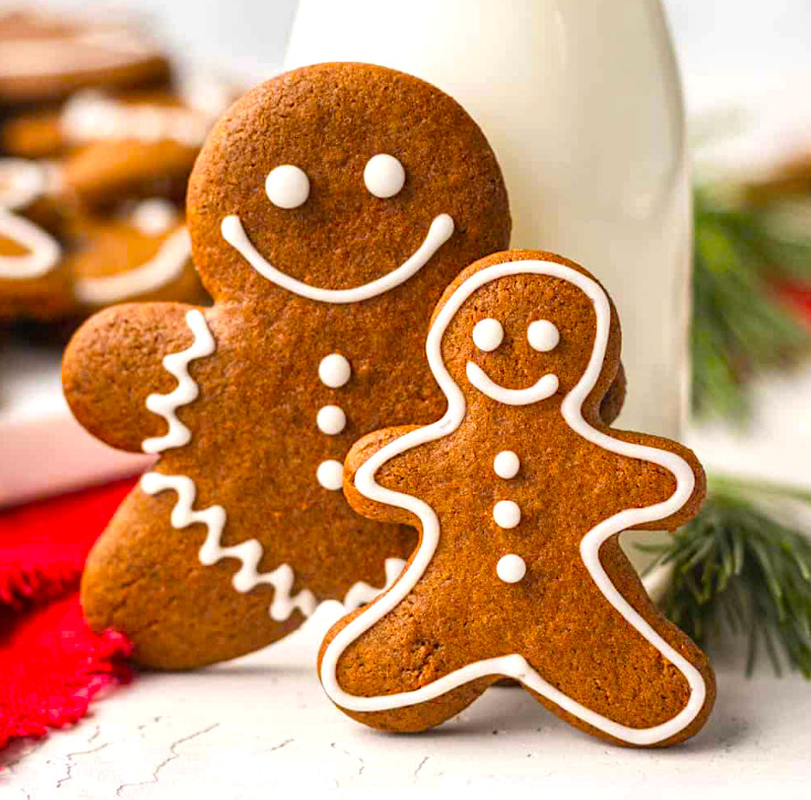 Two smiling gingerbread cookies with white icing decoration next to a glass of milk
