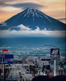 Mount Fuji with snow-capped peak above clouds, urban street in foreground, orange sunset sky
