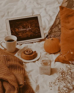 Cozy fall flatlay with tablet, coffee mug, cinnamon roll, candle, pumpkin, and knit sweater on white bedding