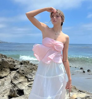 Woman in strapless white and pink bow dress posing on rocky beach with ocean behind her