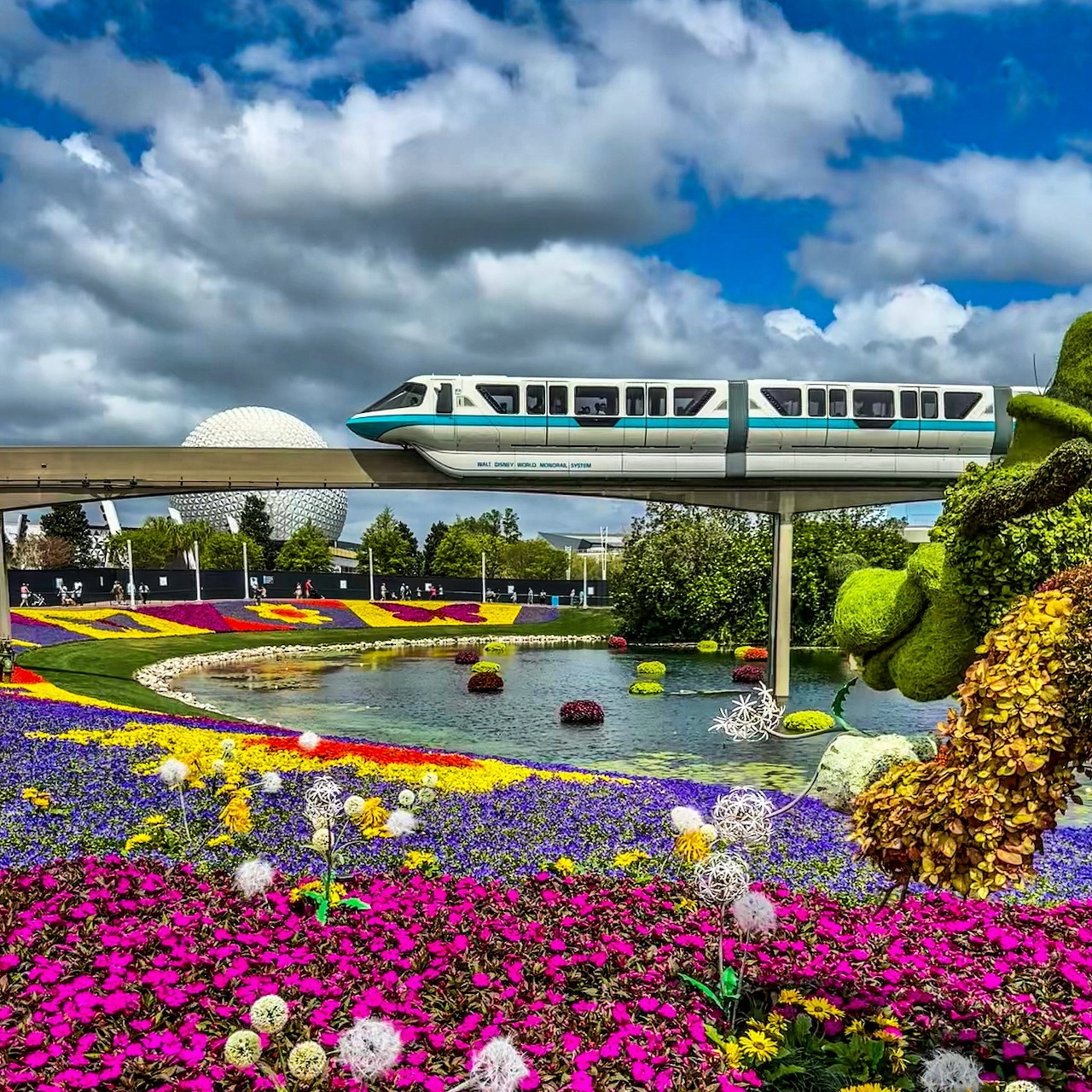 Blue monorail crossing above colorful flower garden with geometric patterns and white dome in background
