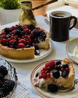 Two berry tarts with raspberries and blackberries on plates beside black coffee mug and brass coffee pot