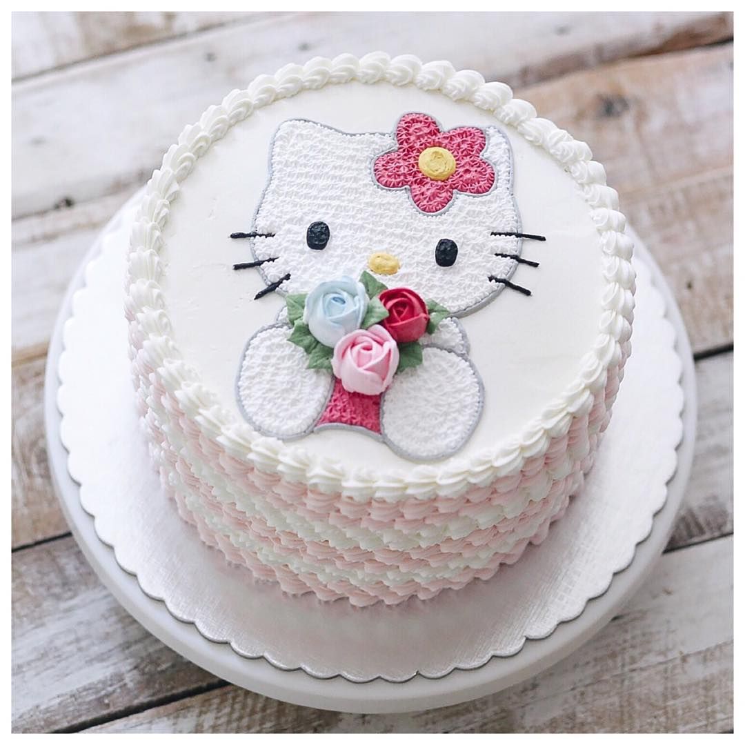 White frosted cake with Hello Kitty face decoration featuring pink bow and rose bouquet on wooden table