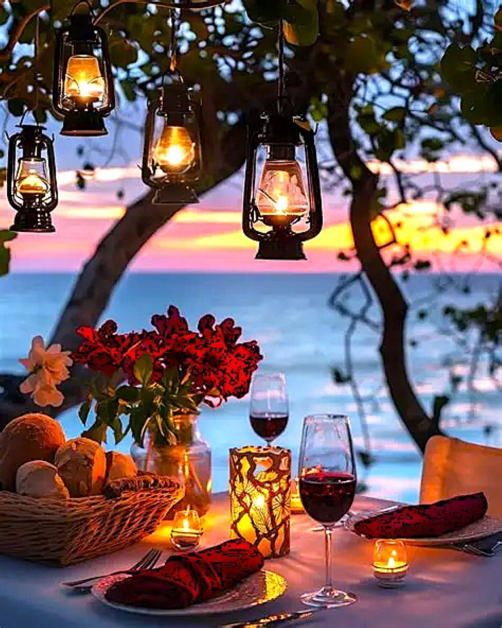 Romantic beach dinner table with lanterns, wine glasses, flowers, and candles at twilight