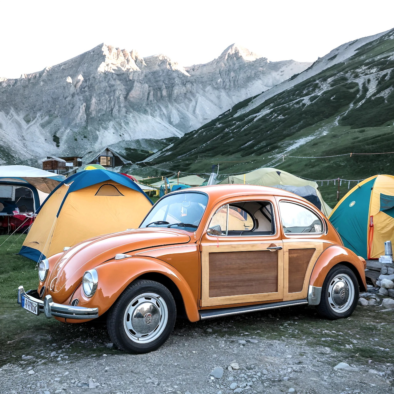Orange VW Beetle with wood trim at mountain camping site with tents and snowy peaks