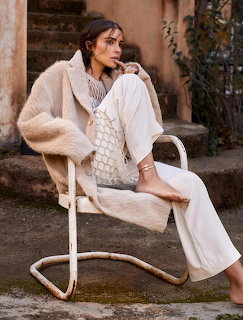 Woman in white outfit with fur vest seated on white cantilever chair by stone wall
