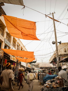 Busy street market with orange tarps overhead, people walking, and shops lining both sides.