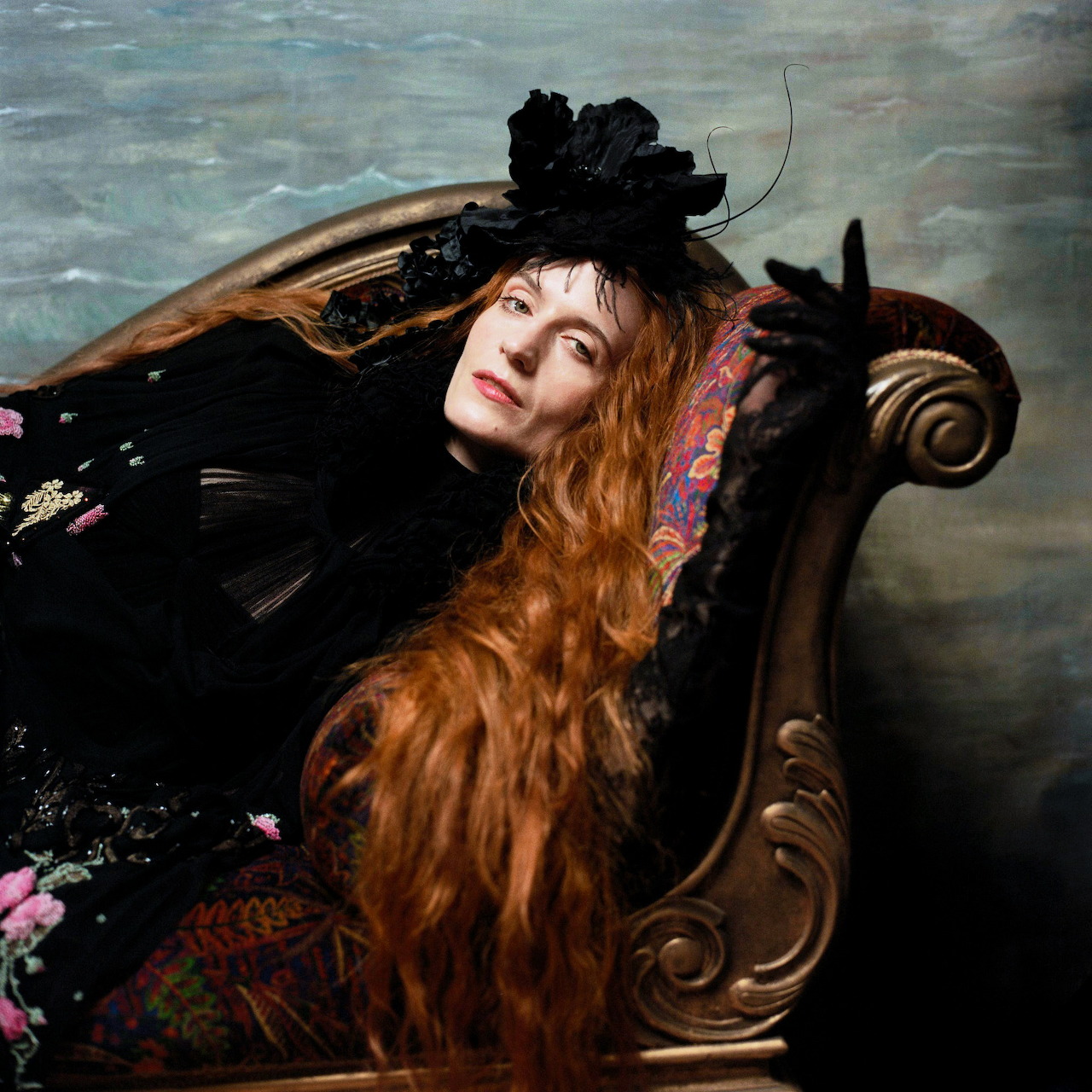 Florence Welch reclining on an ornate chaise lounge with long red hair, black dress, and black fascinator