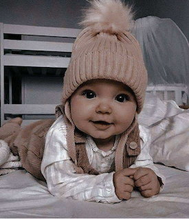 Smiling baby with big eyes wearing beige knit hat with pom-pom, lying on white bedding