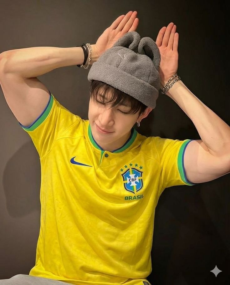 Person in Brazil yellow soccer jersey and gray beanie making playful hand gesture against black background