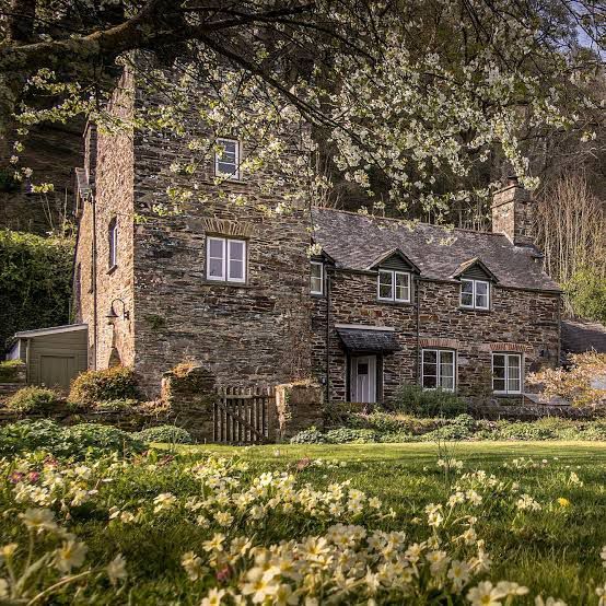 Rustic stone cottage under blooming trees with a grassy lawn and flowers in the foreground