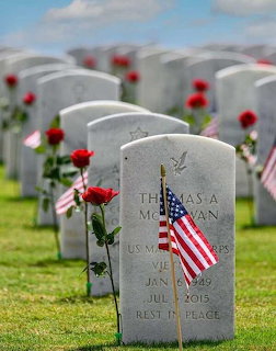 Rows of white military headstones with American flags and red flowers in a cemetery under cloudy sky