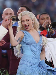 Lady Gaga singing on stage wearing a flowing blue dress with blonde wavy hair