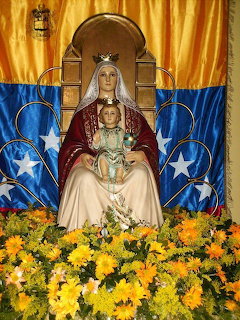 Statue of Our Lady of Coromoto with child Jesus, in front of a yellow and blue flag and yellow flowers