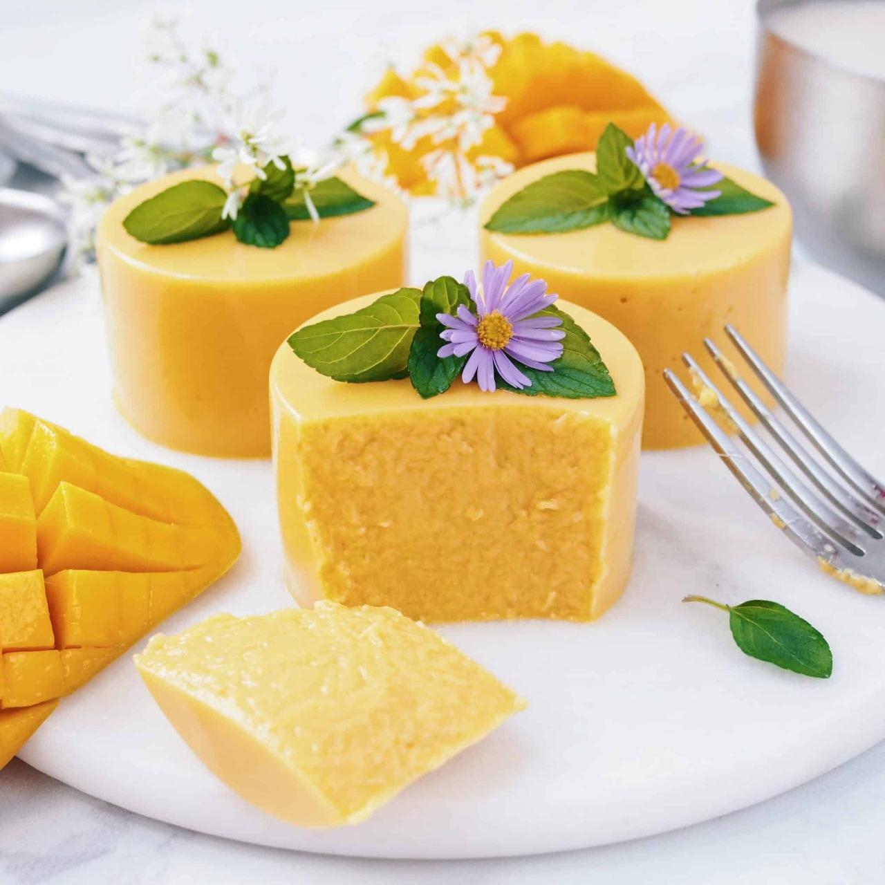 Three small square mango mousse cakes topped with purple flowers and mint on white plate with mango slices