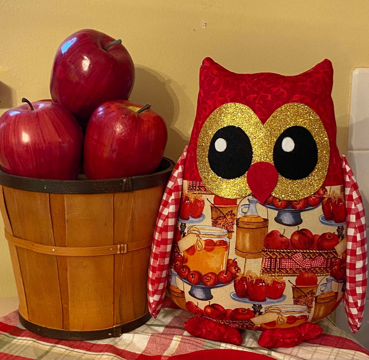 Owl-shaped decorative pillow beside a wooden bucket of red apples on a checkered tablecloth