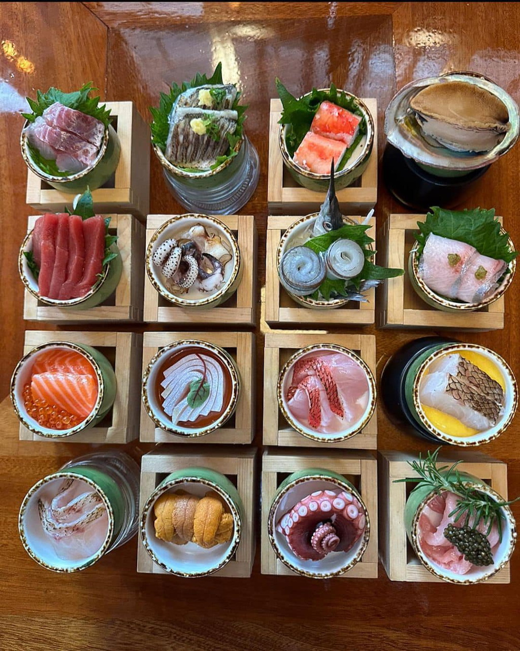 Top-down view of 12 wooden trays each holding a single sashimi portion with various fresh fish