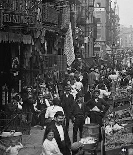 Black-and-white scene of a crowded old New York street with people, carts, and market stalls