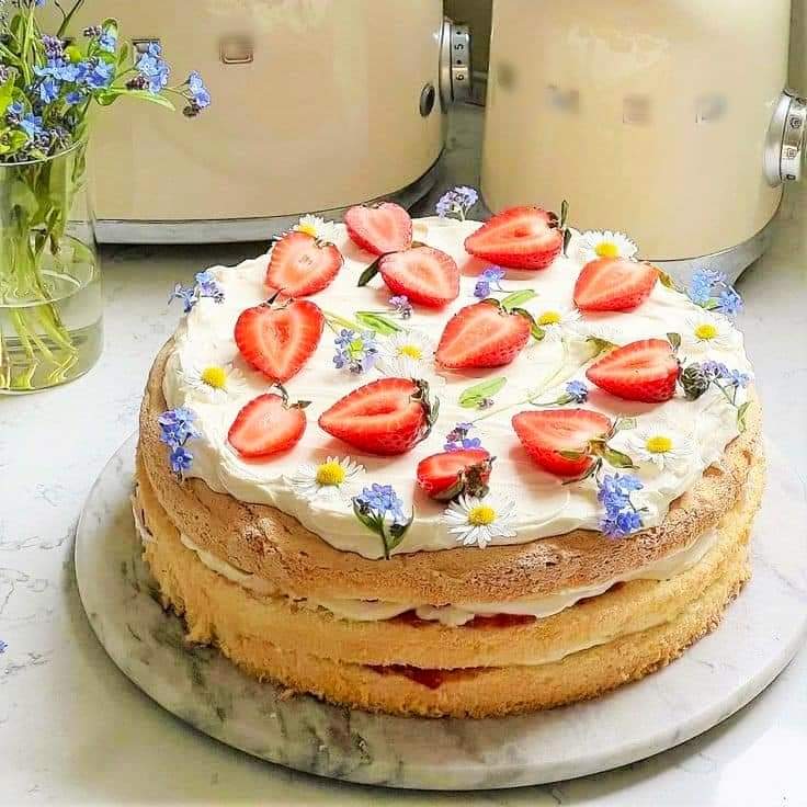 Layered sponge cake with whipped cream, sliced strawberries, and small flowers on a marble stand
