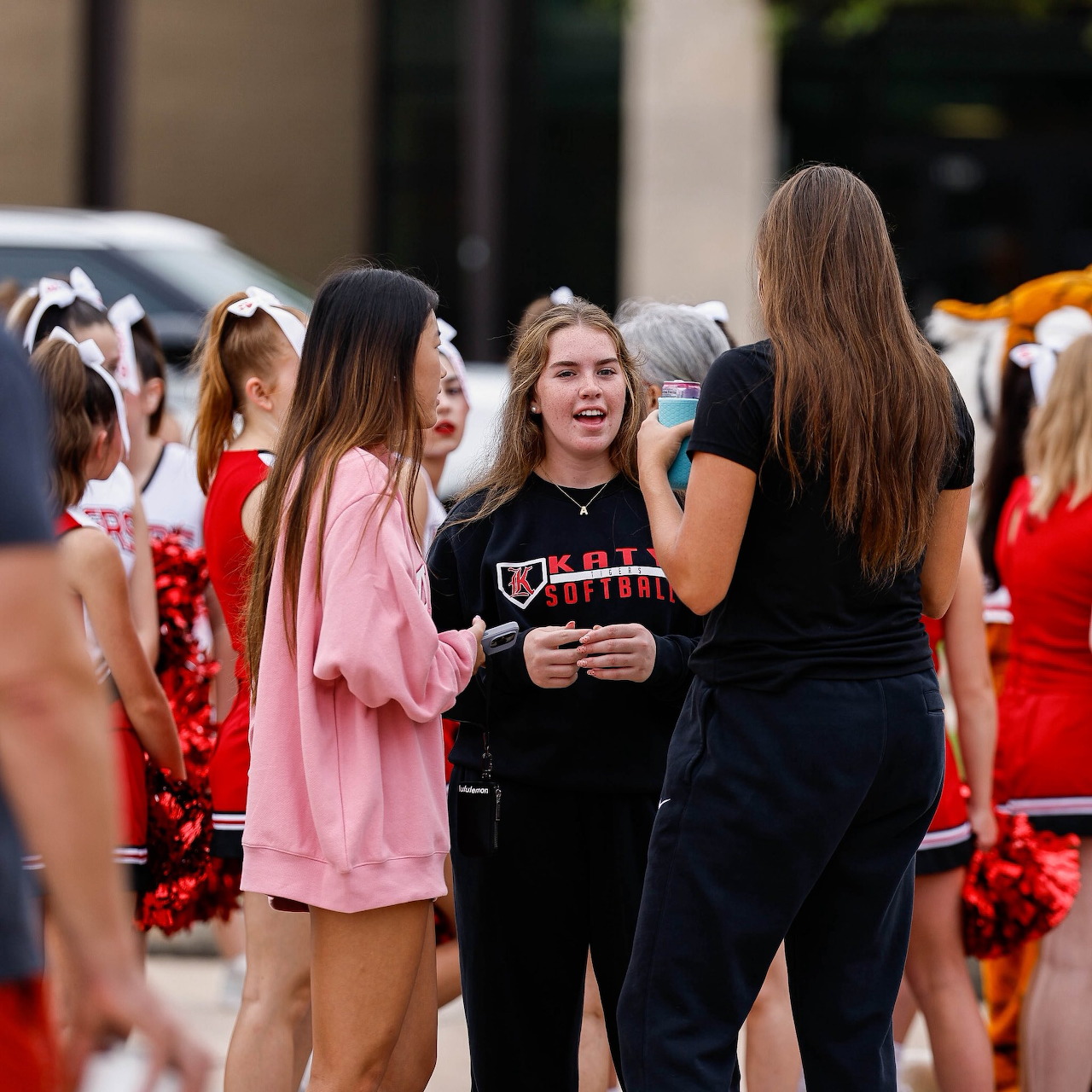 Group of teenage girls talking outdoors near a bus with cheerleaders in red uniforms in the background