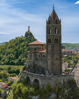 Medieval stone bell tower and chapel on a rocky outcrop with a red statue on a distant peak in Le Puy-en-Velay