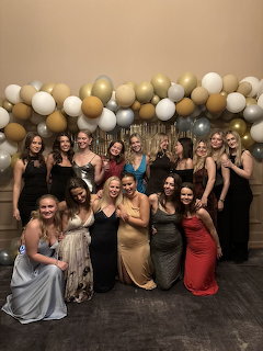 Group of young women in formal dresses posing under a gold and white balloon arch