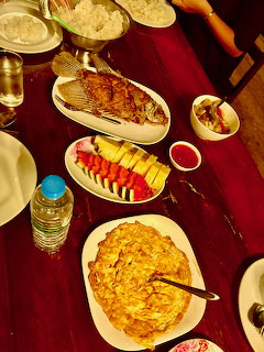 Overhead view of a dinner table with grilled fish, omelet, fruit platter, rice, and drinks