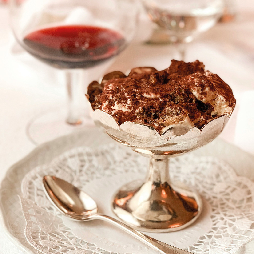 Tiramisu dessert in crystal glass cup on lace doily with spoon and wine glass