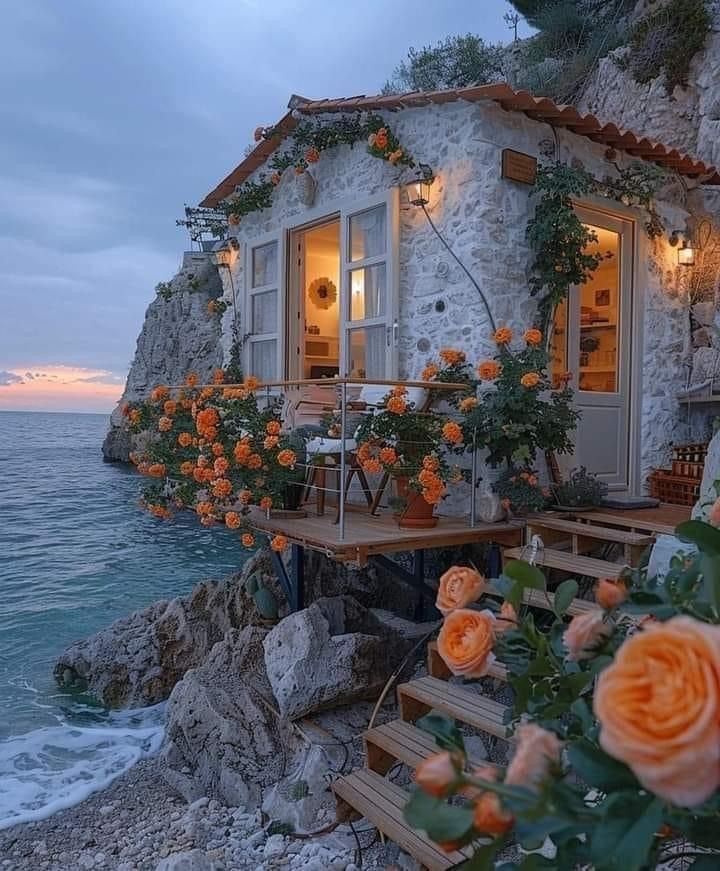 Stone house on cliff edge at sunset with glowing windows and orange roses on terrace overlooking ocean