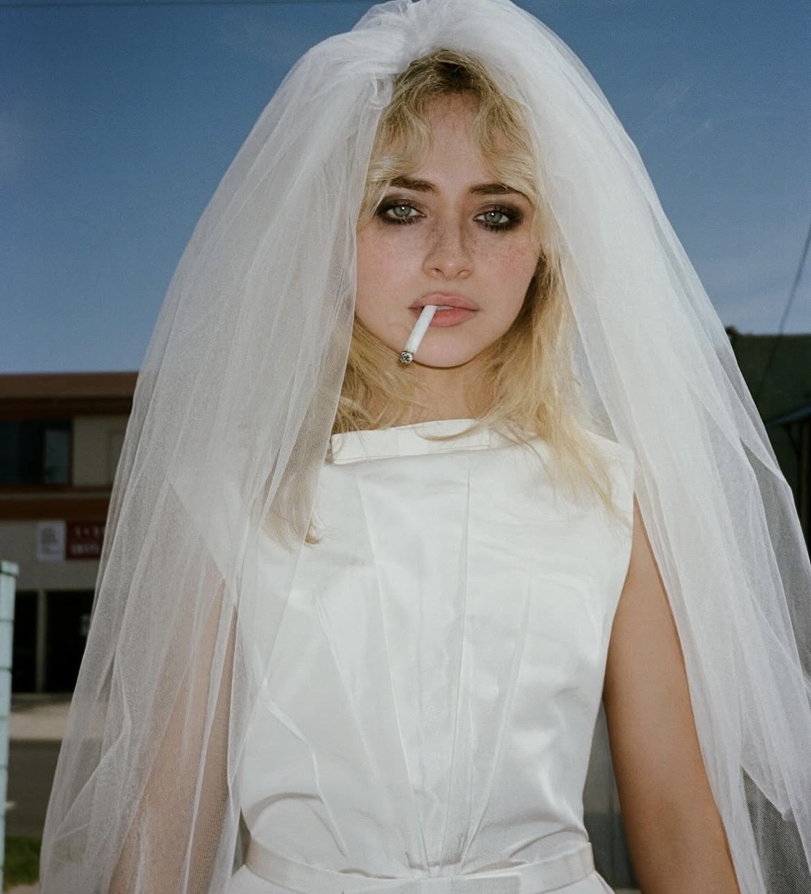 Woman in white bridal dress and long veil outdoors with cigarette against blue sky
