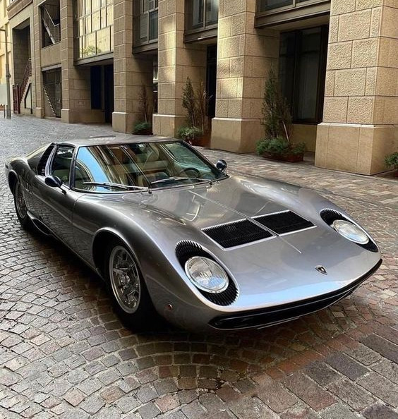 Silver Lamborghini Miura classic supercar parked on cobblestones beside stone buildings