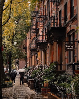 Rainy autumn street with brownstone buildings, fire escapes, fall foliage, and person with umbrella