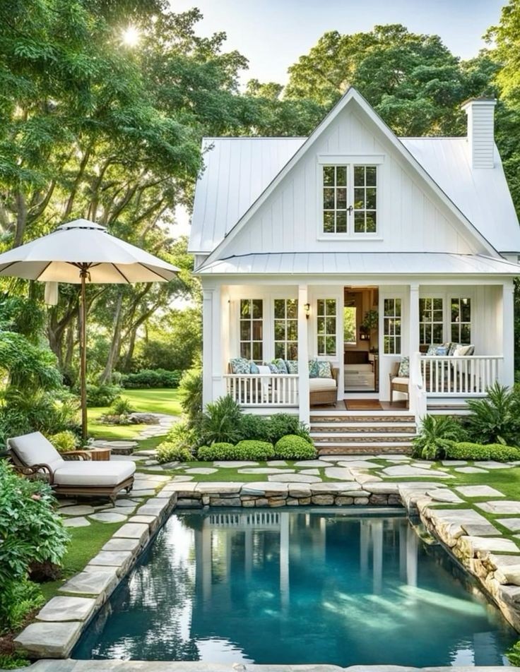 White cottage with porch and rectangular pool surrounded by green trees and garden landscaping