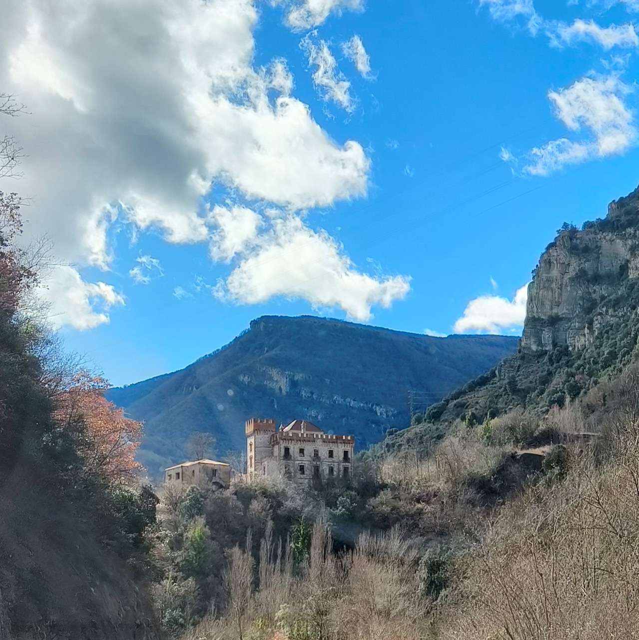 Stone castle among trees with mountain, cliffs, and blue sky with clouds