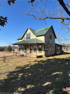 Weathered two-story farmhouse with green roof and porch in rural yard with bare trees